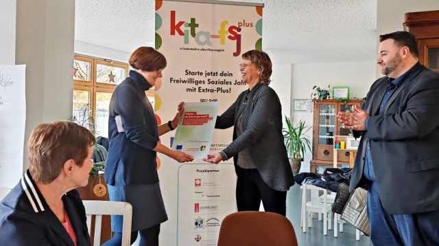 Das Foto zeigt vier Personen, in der Mitte zwei Frauen, die eine Förderplakette miteinander austauschen.